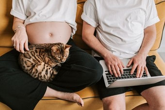 Picture: A pregnant woman sitting with a cat in her lap while her partner is browsing her pregnancy checklist on a laptop